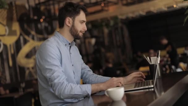 Мужчина с ноутбуком за столом в ресторане или баре, мужчина, работающий на ноутбуке в кафе, молодой хипстер с кофе-брейком в баре, он держит чашку и соединяется с ноутбуком, бизнесмен — стоковое видео