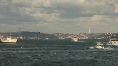Boğaz köprüsünün arka planında tekneler, rüzgarlı hava. Boğaziçi Köprüsü, ön planda turistler ve martılarla dolu eğlence tekneleri..