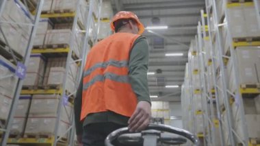 Turuncu yelekli bir adam depoda bir Palet Kamyonu taşıyor.
