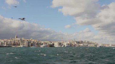Galata Kulesi 'nin yavaş çekim görüntüleri, ön planda turistler ve martılarla dolu eğlence tekneleri. Galata Kulesi 'nin arka planında tekneler, rüzgarlı hava