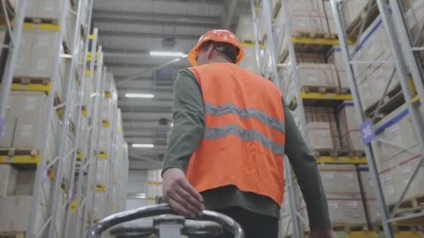 Un homme dans un entrepôt traîne un chariot, L'homme en uniforme et casque marche dans l'entrepôt. — Video