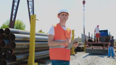 Depo çalışanı metalin envanterini çıkarıyor. Müdür ürünlerin sayısını sayar. Turuncu yelekli bir depo çalışanı kağıda not alır..