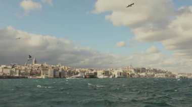 Galata Kulesi 'nin yavaş çekim görüntüleri, ön planda turistler ve martılarla dolu eğlence tekneleri. Galata Kulesi 'nin arka planında tekneler, rüzgarlı hava