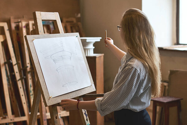 Young beautiful girl student is drawing a Doric capital with a pencil on a wooden easel as her university assignment