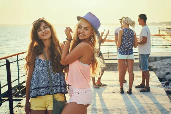 Zwei schöne junge Mädchen haben Spaß am Abend am Meer mit einer Gruppe ihrer Freunde im Hintergrund — Stockfoto