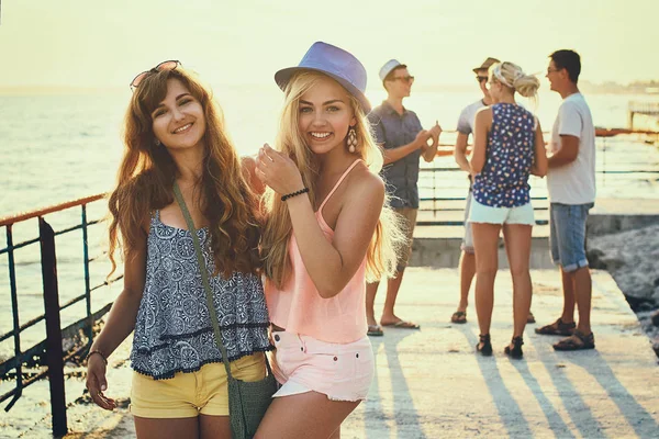 Two beautiful young girls having fun at the evening seaside with group of their friends on background toned in vintage filter style — Stock Photo, Image