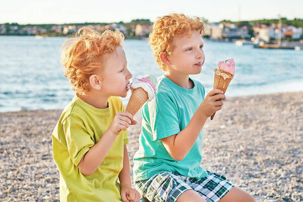 Two cute red curly boys brothers are happy while getting dirty and eating ice cream on the summer beach