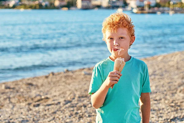 Roztomilý červený kudrnatý chlapec je velmi šťastný, zatímco se špinavé a jíst zmrzlinu na letní pláži — Stock fotografie