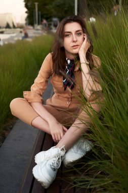 Genç, güzel, kızıl saçlı, genç kız dar giysili kadın bej rengi tulum ya da uçuş kıyafeti giyiyor ve uzun, yeşil bir bankta oturan beyaz, tıknaz bir platform spor ayakkabısı giyiyor.