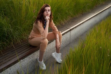 Genç, güzel, kızıl saçlı, genç kız dar giysili kadın bej rengi tulum ya da uçuş kıyafeti giyiyor ve uzun, yeşil bir bankta oturan beyaz, tıknaz bir platform spor ayakkabısı giyiyor.