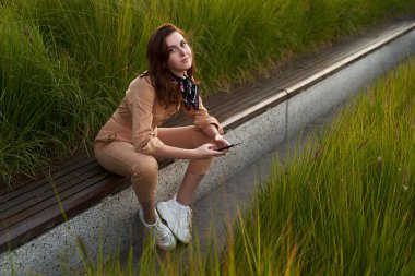 Genç, güzel, kızıl saçlı, genç kız dar giysili kadın bej rengi tulum ya da uçuş kıyafeti giyiyor ve uzun, yeşil bir bankta oturan beyaz, tıknaz bir platform spor ayakkabısı giyiyor.