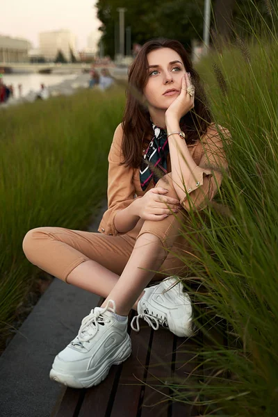 Young beautiful red haired gen z girl wearing slim fitted women beige khaki coverall overall denim jumpsuit or flight suit and white chunky platform sneakers sitting on a long park bench in high green — 스톡 사진