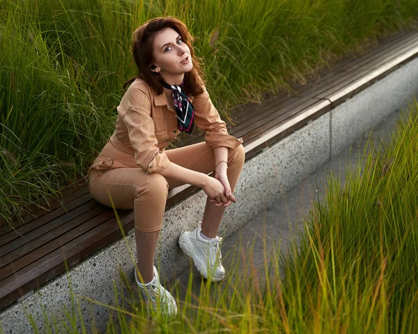 Junge schöne rothaarige Mädchen tragen schlanke taillierte Frauen beige khaki Overall Overall Jeans oder Anzug und weiße klobige Plateau-Turnschuhe sitzen auf einer langen Parkbank in High Green — Stockfoto