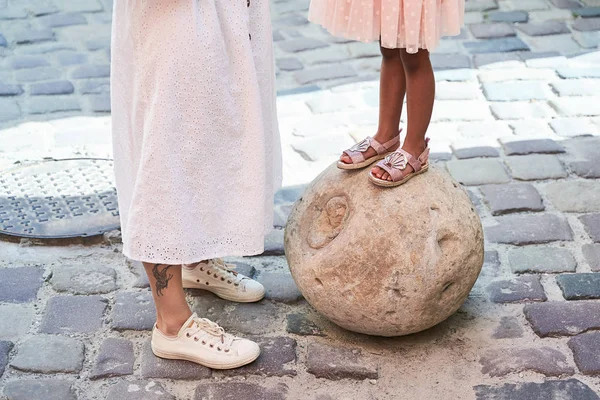 Happy multiethnic family from USA on their European vacation trip. White mother in sneakers and her adorable mixed race daughter in pink dress and sandals standing on a street. Visible legs and feet. — Stock Photo, Image