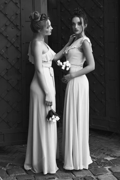 Two beautiful bridesmaids girls blonde and brunette ladies wearing elegant full length light color tulle one shoulder bridesmaid dress and holding flower bouquets. European old town location for — ストック写真