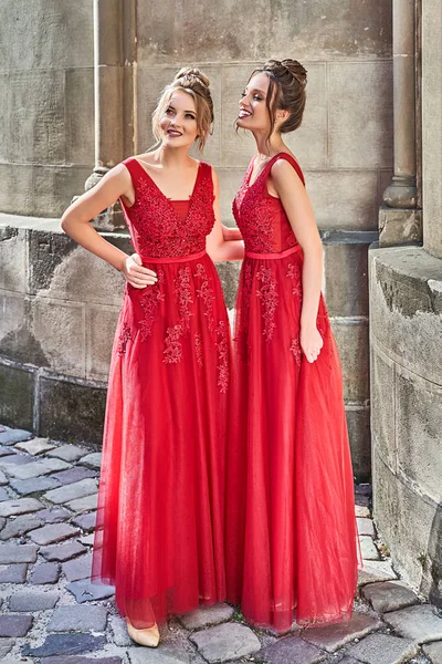Két gyönyörű koszorúslány lányok szőke és barna hölgyek visel elegáns teljes hosszúságú piros sifon koszorúslány ruha csipkével és gazdaság virágcsokrok. Európai óvárosi helyszín az esküvő napján. — Stock Fotó