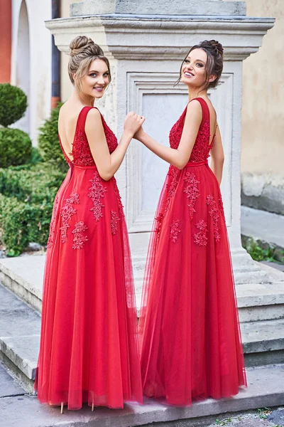 Dos hermosas damas de honor chicas rubias y morenas damas con elegante vestido de dama de honor de gasa roja de longitud completa con encaje y la celebración de ramos de flores. Ubicación del casco antiguo europeo para el día de la boda . — Foto de Stock