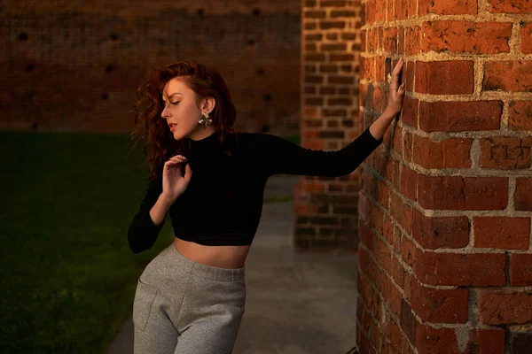 Retrato de uma linda menina ruiva sensual posando ao pôr-do-sol usando luz cinza claro perna larga calças de cintura alta jersey e t-shirt manga longa preta lançando uma sombra na parede de tijolo vermelho — Fotografia de Stock