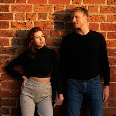 Romantik çift genç, güzel, şehvetli, kızıl saçlı kadın ve uzun boylu, sarışın adam gün batımında kırmızı tuğla duvarda güneş ışığında poz veriyorlar.