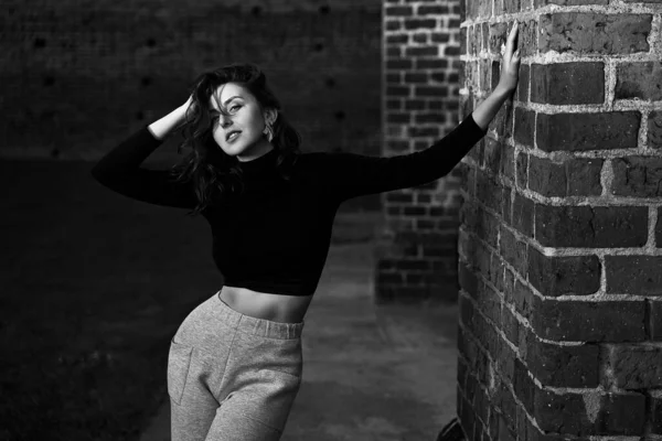 Retrato de una hermosa chica pelirroja sensual posando en la luz del sol puesta del sol usando pantalones de jersey de cintura alta de pierna ancha gris claro y camiseta negra de manga larga que proyecta una sombra sobre el fondo de la pared de ladrillo en — Foto de Stock