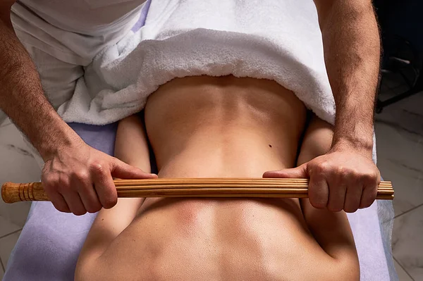 Muž masér dělá japonské masáže s bambusovými tyčinkami na krásném nahém ženském těle v lázeňském salonu. Koncept kosmetické léčby. — Stock fotografie