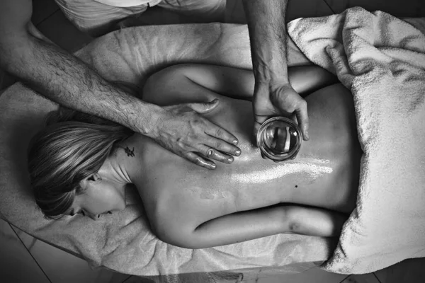 Mužský masér provádí masáže pomocí speciálního aromatického oleje na krásném nahém ženském těle v lázeňském salonu. Koncepce kosmetické léčby v černé a bílé. — Stock fotografie