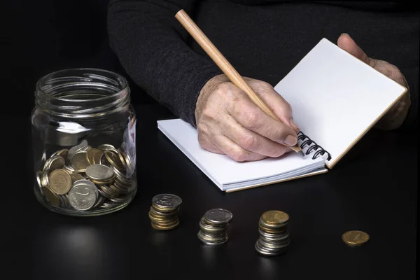 The hands of an elderly woman writing down their expenses in a notebook. A jar of small coins on a dark background.