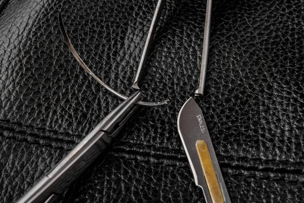 Needle in needle holder scalpel surgical tweezers on a black leather texture background