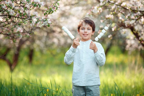 Lycklig pojke med marshmallow på pinnar i blommande trädgård — Stockfoto