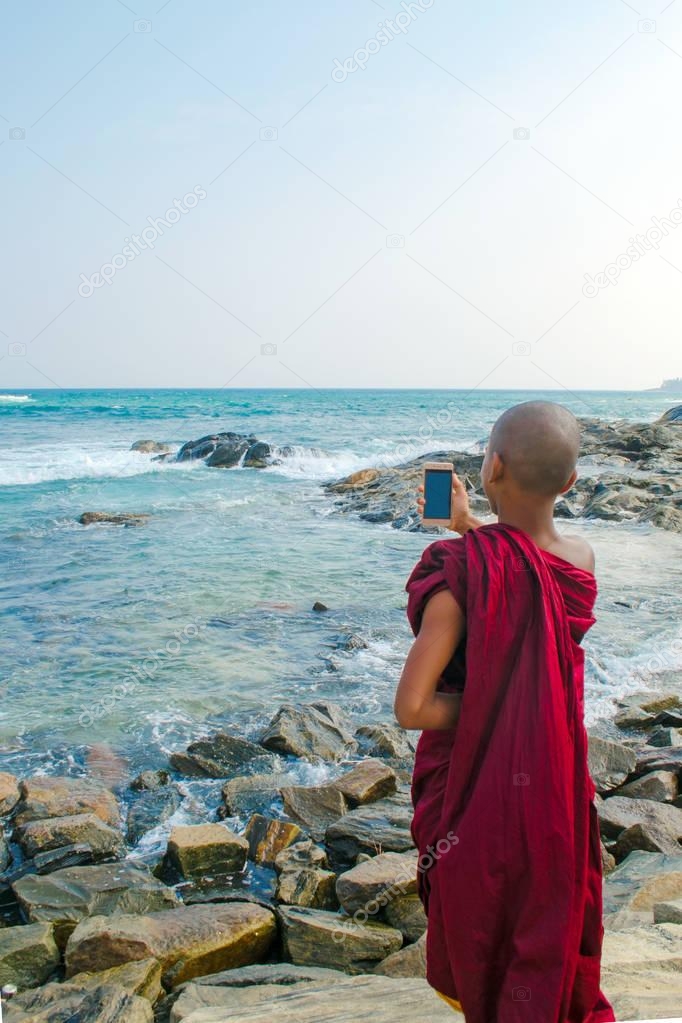 Buddhist monk holds a mobile phone on the ocean beach