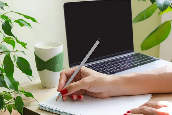 Una donna con manicure brillante scrive a matita nel diario. Accanto al tavolo c'è un computer portatile, una tazza d'acqua con limone e piante verdi. Il concetto di un luogo di lavoro o di un ufficio ecologico . — Foto Stock