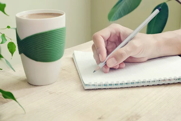 A mão de uma mulher com um lápis é escrita em um diário com espirais. Ao lado da mesa está uma xícara de café e planta com folhas verdes. Vista lateral — Fotografia de Stock