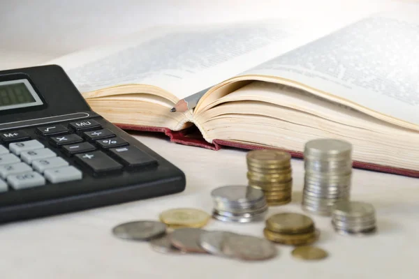 Monete sfocate in pile e una calcolatrice sullo sfondo di un libro aperto con una matita. Il concetto di costi elevati dell'istruzione per gli abitanti dei paesi poveri — Foto Stock