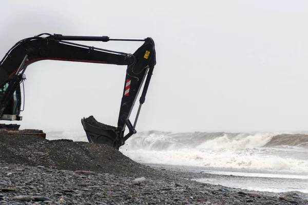 The excavator works with a bucket full of stones on the seashore in stormy weather.