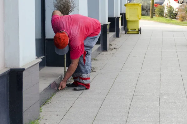 Rengöring av en stadsgata - arbetaren gården djurhållaren i uniform samlar sopor av händerna nära en byggnad, bakom det där är en papperskorg. — Stockfoto