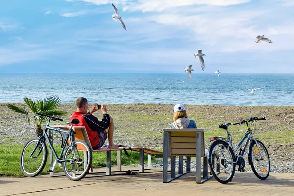 Rest after weekend bike trip - a couple sit on deckchairs and enjoy a beautiful view of the sea and seagulls, making photo on the smartphone.