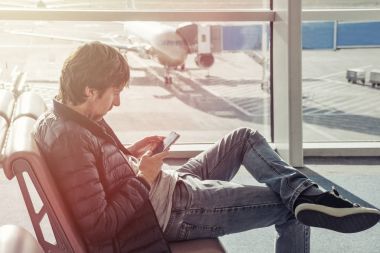  Kot pantolon ve ceket oturur sandalyeye genç adam Havaalanı lounge alanında cep telefonu kullanarak zaman harcamak. Online bankacılık gelmeden yabancı ülkede otel rezervasyon. Arka plan, jetway ile uçak.
