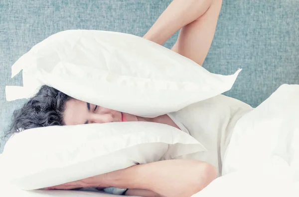 Young caucasian woman with black hair sleeps covering her head and ears with pillows.