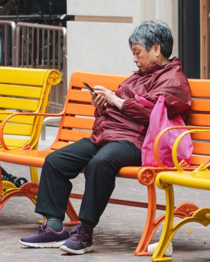 Asyalı yaşlı kadın şehirdeki bir bankta akıllı telefon kullanıyor. Hong Kong, 2018-03-06 