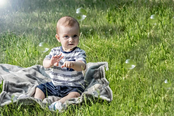 Een schattige baby zitten en spelen met zeepbellen in de natuur in een park van de zomer op gras — Stockfoto