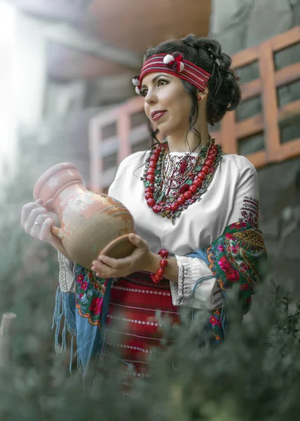 Una mujer muy joven vestida con un traje popular ucraniano sosteniendo una jarra de barro en el patio. Cosplay. —  Fotos de Stock