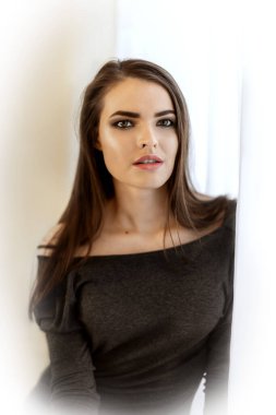 Close-up portrait of young pretty female 20-30 years old dressed in a feminine dress with open neckline standing next to window with white curtains and looking into camera