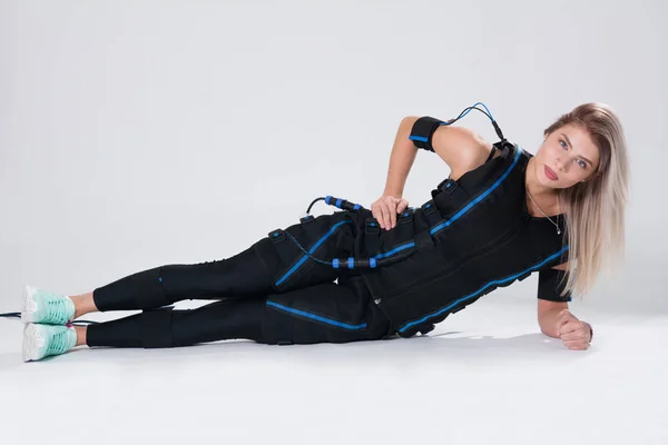 Blonde in an electric muscular suit for stimulation makes an exercise on the rug. — Stock Photo, Image