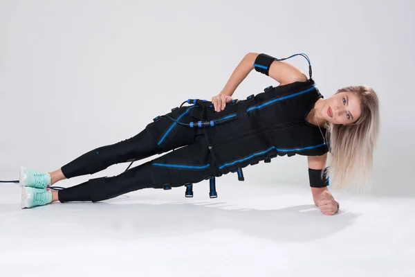Beautiful blonde in an electric muscular suit for stimulation makes an exercise on the rug — Stock Photo, Image
