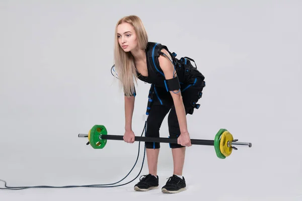 Beautiful blonde in an electric muscular suit for stimulating exercises with a barbell — Stock Photo, Image