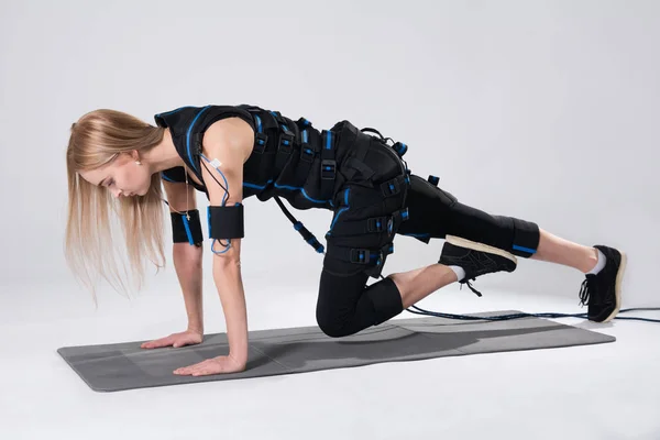 Loira em um terno muscular elétrico para estimulação faz um exercício no tapete . — Fotografia de Stock