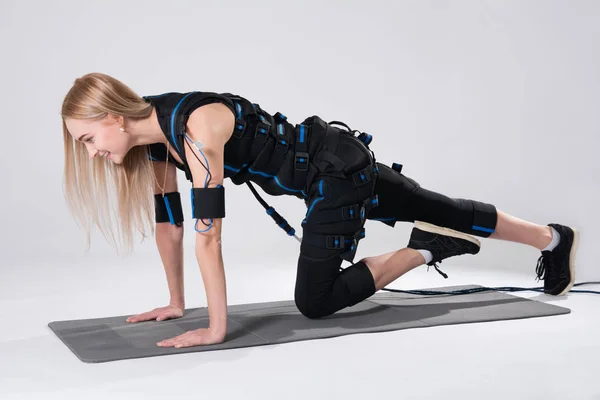 Hermosa rubia en un traje muscular eléctrico para la estimulación hace un ejercicio sobre la alfombra — Foto de Stock