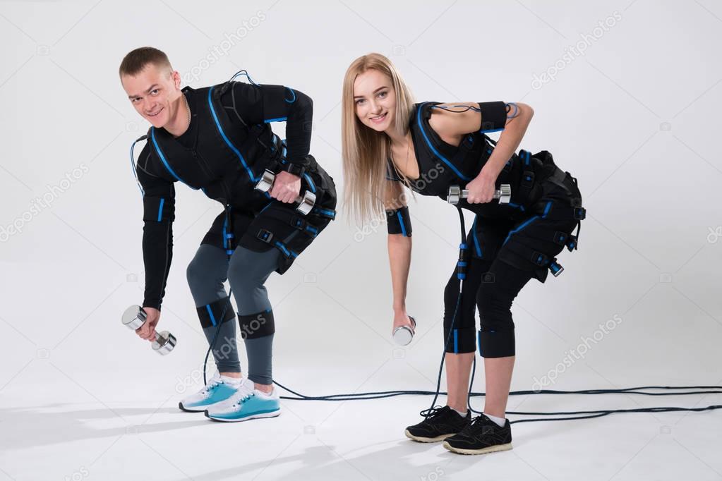 Young man and woman in electric muscular suit for stimulation with dumbbells