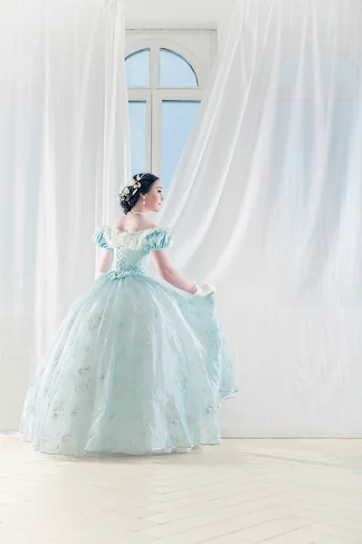 Elegant woman at a high window in historic dress, straightens the curtains, and waiting for friends to prom to dance