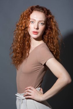 Ciddi kızıl saçlı kıvırcık saçlı kadın, doğrudan kameraya bakıyor, ellerini kemerinde tutuyor. İzole edilmiş gri bir arkaplanda